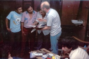 Man with white shirt on holds photos up and talks to three other men in an office setting.